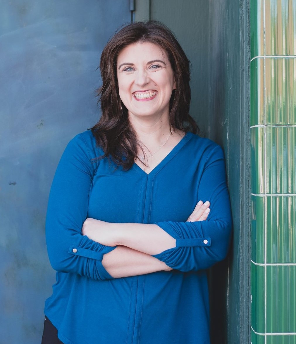 Heather in a blue shirt leaning against a blue door frame, smiling at the camera with arms folded